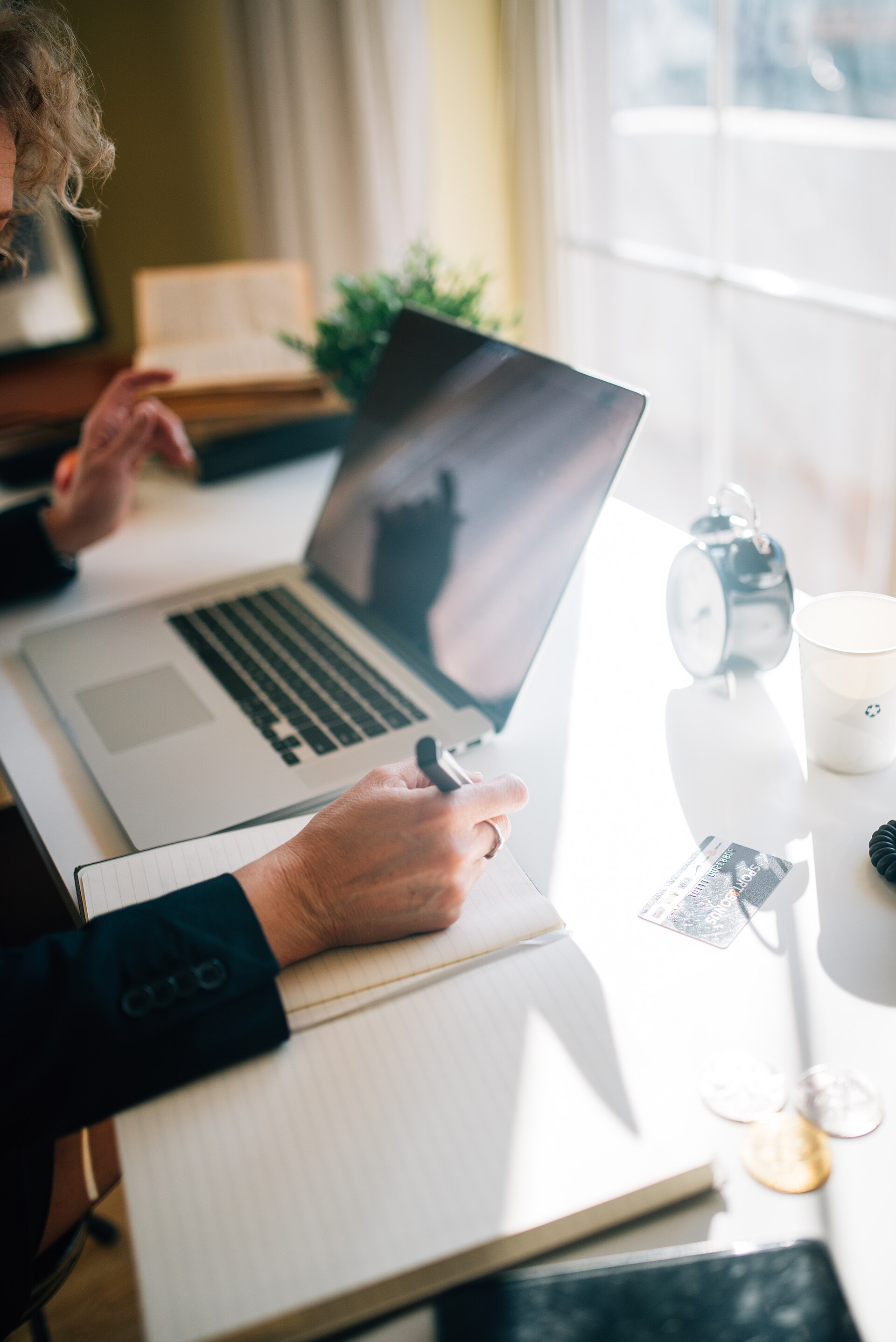 Person writing notes while working on a laptop at a bright desk